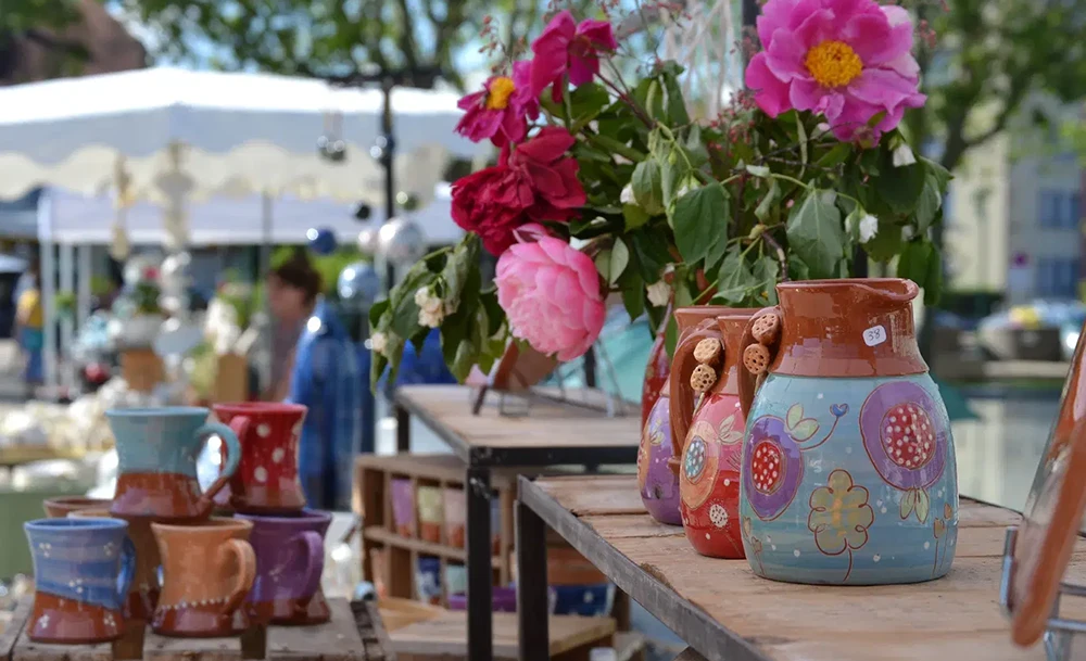 Poterie colorée au Marché potier Rumilly, avec des jarres décorées de motifs floraux et des fleurs vibrantes en arrière-plan.
